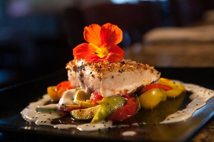 Crusted fish fillet topped with an edible orange flower, served over colorful vegetables
